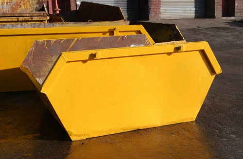 A large, yellow industrial skip with a rectangular shape and reinforced edges, positioned on a dark paved surface in an outdoor area. The skip's sides show scratches and signs of weathering, indicating frequent use. Behind it, there are additional similar skips and a section of a warehouse or storage facility with metal roll-up doors. The lighting appears natural, casting subtle shadows on the ground and highlighting the vivid yellow finish of the skip. This type of skip is typically used for waste collection and rubbish removal in commercial or construction settings, aligning with the services provided by Commercial Waste Richmond upon Thames in the local postcode district. The environment suggests an industrial or waste management site within the Richmond upon Thames area, suitable for handling large quantities of waste material.