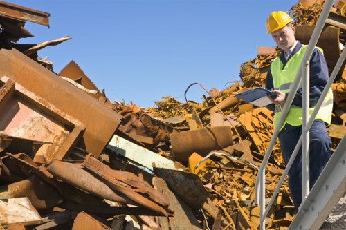 A man in a yellow safety helmet and high-visibility vest stands on a metal staircase at a rubbish disposal site, holding a clipboard and examining a large pile of rusted metal scraps. The debris consists of various distorted and weathered metal sheets, beams, and structural components, with textures showing rust, corrosion, and peeling paint in shades of brown, orange, and yellow. The background features a clear blue sky, indicating a bright and sunny day, while the surrounding environment suggests an industrial or waste management area. The scene relates to commercial waste removal services, particularly in Richmond upon Thames, with the presence of rusted scrap metal highlighting the type of rubbish that might be collected and processed by Commercial Waste Richmond upon Thames.