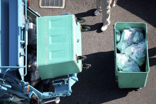 An overhead view of a green waste collection bin filled with plastic bags containing rubbish, positioned on a paved surface outdoors. To the left, part of a blue commercial waste container or vehicle is visible, indicating a waste collection or rubbish removal process. In the background, a person wearing light-colored clothing and white shoes stands nearby, slightly out of focus, suggesting an ongoing clearance or disposal activity. The setting appears to be an industrial or commercial area, consistent with waste management services in Richmond upon Thames. The image emphasizes the handling of general rubbish within a waste collection context, aligning with services offered by Commercial Waste Richmond upon Thames for rubbish removal in the local area.