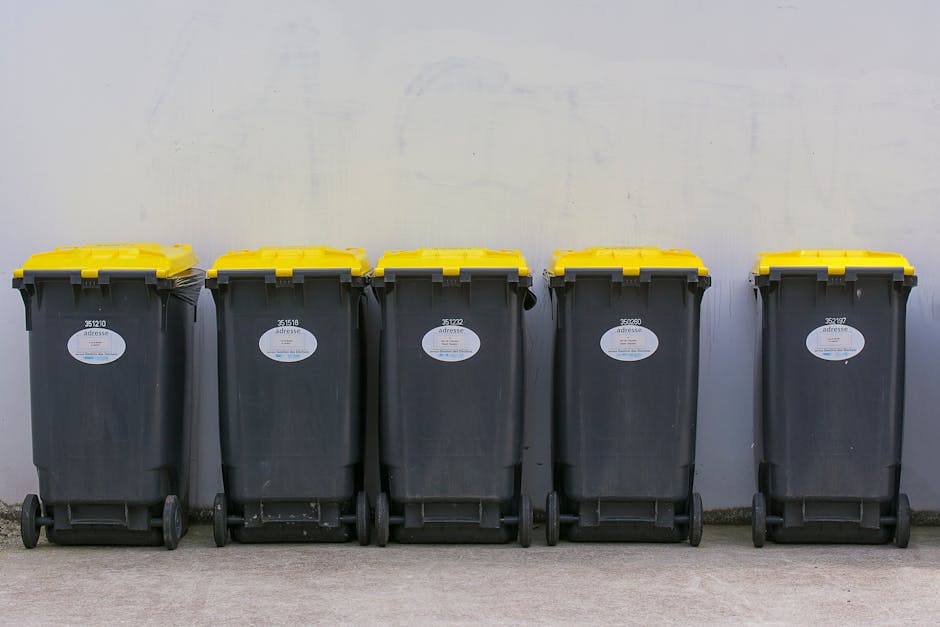 A pile of discarded cardboard boxes, some flattened and others partially assembled, is situated against a weathered brick wall and a rough concrete block wall under natural daylight. The boxes vary in