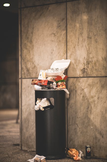 An outdoor urban scene shows a large, rusted metal waste container painted in faded red, positioned against a dark green wall on the left and a light grey textured wall on the right. The container is 