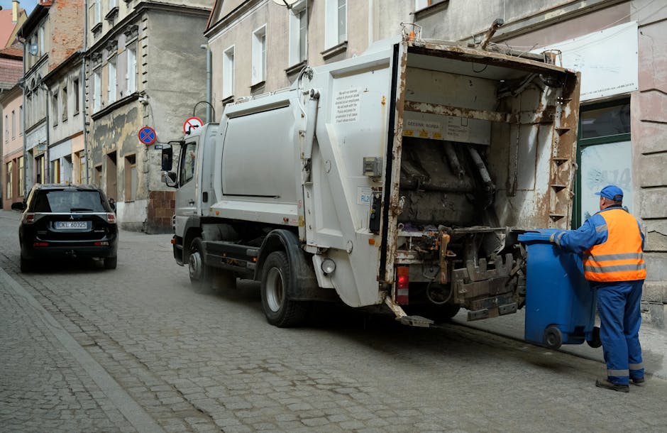 A pair of large black trash bags filled with waste material are positioned on the edge of a paved street or sidewalk in front of a black metal fence with vertical bars. The bags appear to be made of d